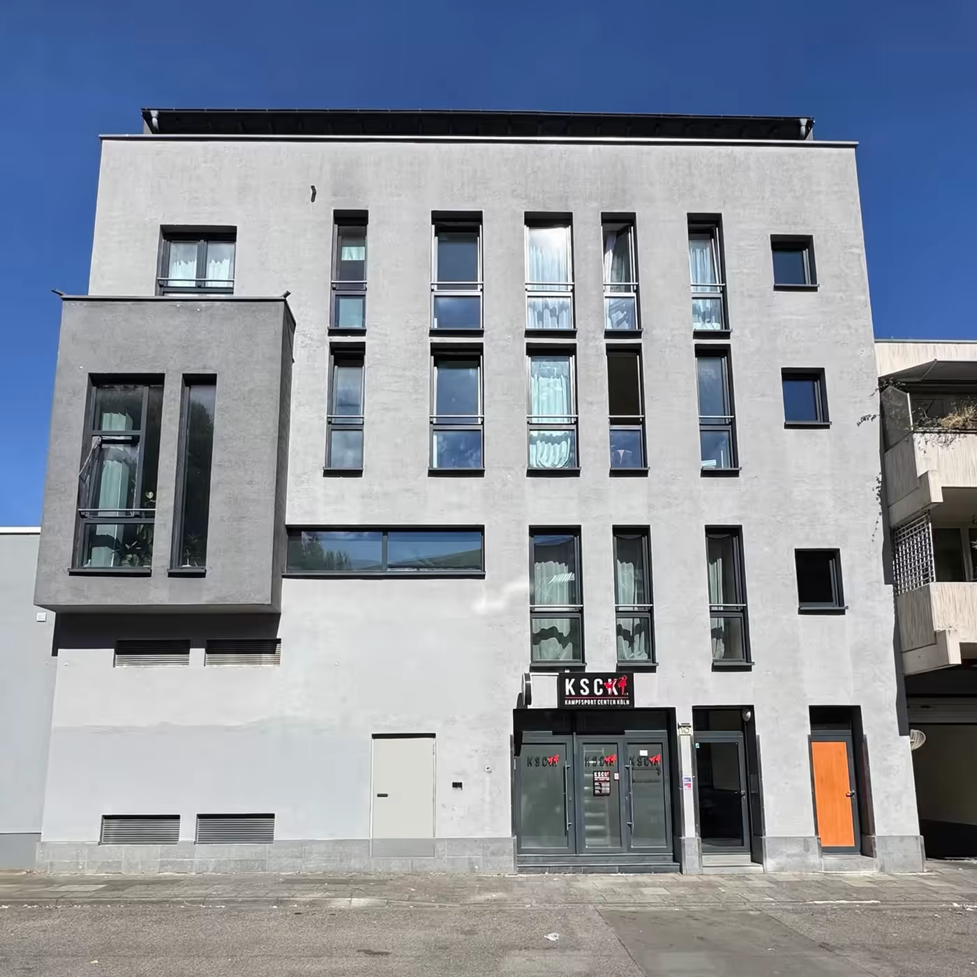 Heinrich-Brüning-Straße 10 in Köln, Straßenansicht: fünfgeschossiges Wohn- und Geschäftshaus mit hellgrauem Putz, dunklen Fensterrahmen und vorspringendem Erker.
