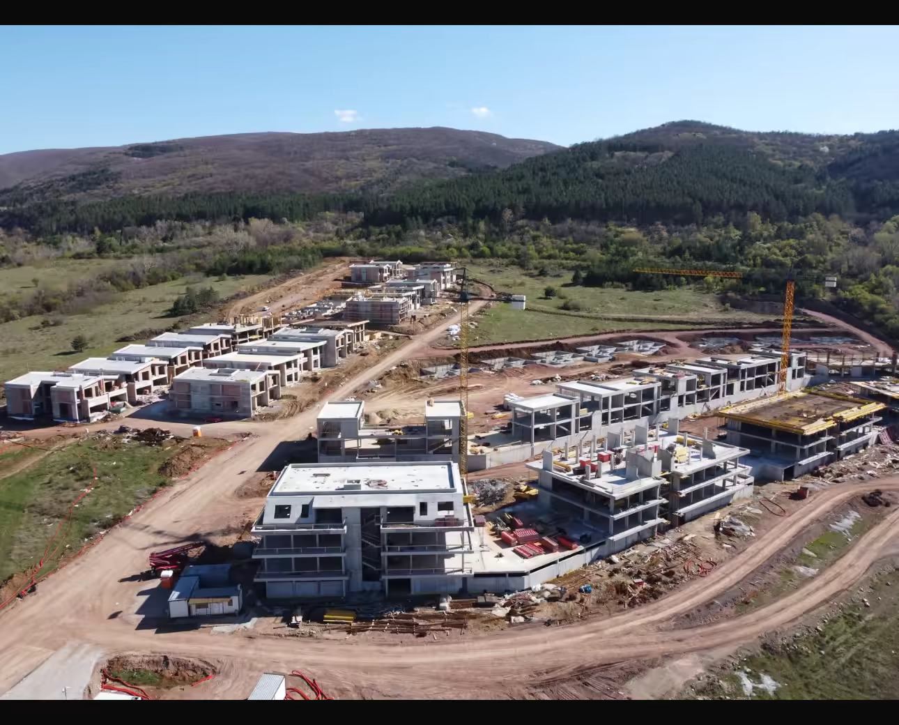 Aerial view of the Newland District construction site near Sofia: terraced houses and apartment buildings in shell construction, two tower cranes, wooded slopes behind.