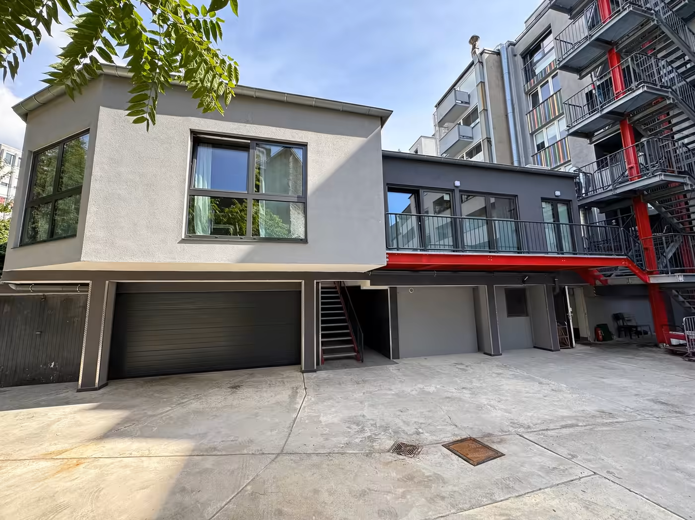 Neubau im Innenhof der ZÜL 6 in Köln: zweigeschossiger, hellgrau verputzter Baukörper mit großen Fenstern über einem Garagentor, rechts ein dunkler Anbau mit Balkon auf roten Stahlträgern und einer Außentreppe.